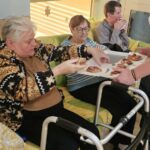 Osebe sedijo na kavču v skupnem prostoru, ena oseba prejema kos kruha z oblogo s pladnja, ki ga drži druga oseba.