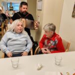 Tri osebe so zbrane ob mizi v skupnem prostoru, ena oseba stoji in igra na harmoniko, dve osebi sedita in poslušata.