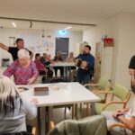 Več oseb je zbranih v skupnem prostoru okoli miz, ena oseba stoji in igra na harmoniko, druge sedijo ali stojijo ter spremljajo dogajanje.