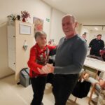 Dve osebi stojita v skupnem prostoru in se držita za roke, kot pri plesu, v ozadju so mize in druge osebe.