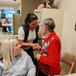 Ena oseba stoji ob mizi in se rokuje z drugo osebo, ob njiju sedi še ena oseba, na mizi je krožnik z ostanki sladice.