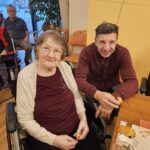 Dve osebi sedita ob mizi v skupnem prostoru in držita koščke peciva, v ozadju so vidne druge sedeče osebe.