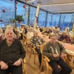 Več oseb sedi za mizami v večjem skupnem prostoru in posluša dogajanje, na mizah so prtički in pecivo, prostor je praznično okrašen.