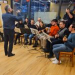 Skupina oseb sedi v polkrogu z glasbili in notnimi stojali, ena oseba stoji pred njimi in z gibi rok vodi nastop.