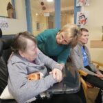 Oseba se prijazno skloni k stanovalki, ki sedi na invalidskem vozičku, v skupnem prostoru doma.