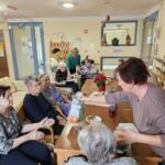 Skupina stanovalcev sedi okoli mize v skupnem prostoru, kjer poteka druženje in razdeljevanje daril.