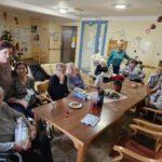 Skupina stanovalcev, zbranih okoli mize v skupnem prostoru doma, ob prazničnem druženju.