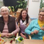 Tri osebe sedijo za mizo, dve osebi držita rdeče nageljne, na mizi so kozarci s pijačo in prtički.