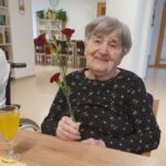 Oseba sedi za mizo in drži šopek rdečih nageljnov, na mizi je kozarec z rumeno pijačo.