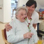 Ena oseba sedi in drži rdeče nageljne, druga oseba stoji za njo in jo drži okoli ramen, na mizi je kozarec s pijačo.