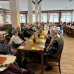 Več oseb sedi ob dolgih mizah v jedilnici, pred njimi so kozarci pijače in cvetje, osebe se pogovarjajo.