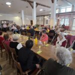 Skupina oseb sedi ob mizah v večnamenskem prostoru in gleda proti skupini otrok, ki stojijo spredaj.