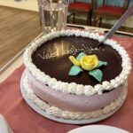 Na vozičku je torta s čokoladno glazuro in okrasno rožo, vanjo je usmerjen nož za rezanje.