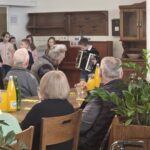 Otrok stoji pred klavirjem in igra harmoniko, v ozadju stojijo drugi otroci, odrasli sedijo in opazujejo.