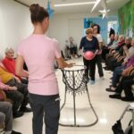 Oseba stoji ob košu z mrežo in podaja žogo, druge osebe sedijo ob stenah in opazujejo igro v hodniku.