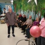 Oseba hodi po prostoru mimo sedečih oseb, v ospredju je žoga, v ozadju je stenska poslikava dreves.