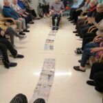 Več oseb sedi v krogu na stolih, na tleh so razporejeni časopisni listi, ena oseba sedi na sredini kroga.