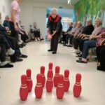 Oseba meče žogo proti rdečim kegljem, druge osebe sedijo ob straneh in spremljajo dogajanje v prostoru.