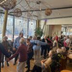 Več oseb se giblje in stoji v večjem skupnem prostoru, nekatere se držijo za roke, druge sedijo ob mizah ali na invalidskih vozičkih.