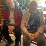 Dve osebi sedita na stolih v notranjem prostoru, ena ima ob sebi pripomoček za hojo, druga pa v rokah drži kozarec z napitkom.