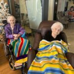 Dve osebi sedita vsaka na svojem sedežu, pokriti z odejama, v prostoru z okrašeno smrečico in steklenimi stenami.