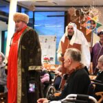 Tri osebe v kostumih stojijo v skupnem prostoru in nastopajo pred sedečimi osebami, okoli so mize, stoli in praznična dekoracija.