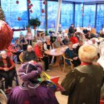 Več oseb sedi za mizami v večjem, praznično okrašenem prostoru in spremlja nastop oseb v kostumih, ki stojijo v ospredju.