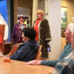 Več oseb sedi za mizo z napitki in opazuje tri osebe v kostumih, ki stojijo in nastopajo v prostoru.