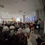 Več oseb sedi na stolih v večnamenskem prostoru in spremlja pevski nastop skupine, ki stoji spredaj.