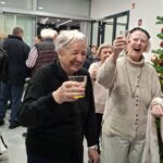 Dve osebi stojita ob okrašeni jelki, se smejita in dvigujeta kozarca s pijačo, v ozadju so druge osebe.