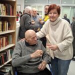 Oseba stoji ob osebi na invalidskem vozičku in drži kozarec pijače, v ozadju so police s knjigami in druge osebe.