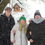 Tri osebe stojijo skupaj na prostem pod drevesom z nameščeno ptičjo krmilnico, okoli njih pada sneg.