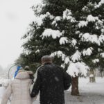 Več oseb hodi po zasneženi poti v parku, ob poti so klopi in veliko zasneženo drevo.