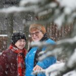 Dve osebi stojita na prostem med sneženjem, oblečeni v zimska oblačila, v ospredju so zasnežene veje drevesa.