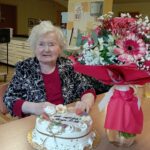 Ženska sedi za mizo s torto in šopkom rož v notranjem prostoru.