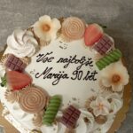 Okrašena torta z napisom stoji na podlagi, okoli so sladki okraski in sadje.