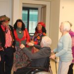 Več oseb v barvitih kostumih stoji ob osebi v invalidskem vozičku in z njo komunicira v notranjem prostoru.