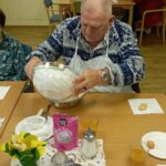 Moški z predpasnikom za mizo preliva sestavine iz posode v skledo, na mizi so sladkor, napitki in krožniki.