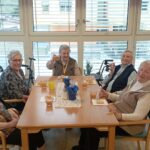 Skupina žensk sedi za mizo ob oknu, pred njimi so kozarci s pijačo in krožniki s pecivom.