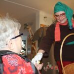 Oseba v prazničnem kostumu podaja majhen predmet sedeči osebi na invalidskem vozičku v notranjem prostoru.