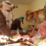 Oseba v kostumu se rokuje s sedečo osebo ob postelji, medtem ko druga oseba v ozadju pripravlja prostor.