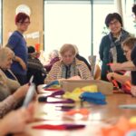 Več oseb sedi za mizo in iz barvnega materiala izdeluje okraske, medtem ko druge osebe stojijo ob njih.