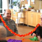 Dve osebi držita dolg verižast trak iz barvnega papirja nad mizo v skupnem prostoru.