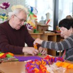 Dve osebi sedita za mizo in skupaj oblikujeta okrasek iz barvnega papirja.