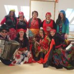 Skupinska fotografija nastopajočih v pisanih kostumih; moški z harmoniko kleči spredaj, ostali stojijo v ozadju v prostoru doma.