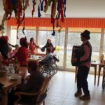 Nastopajoči obiskujejo jedilnico doma; ob mizah sedijo stanovalci, harmonikar stoji ob strani, v ozadju so okna z razgledom na drevesa.