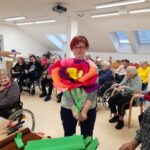 Ženska stoji v prostoru in v rokah drži veliko barvno papirnato rožo, v ozadju sedijo druge osebe.