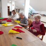 Tri ženske sedijo za mizo in iz barvnega papirja izdelujejo dele dekorativnih rož.