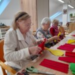 Tri ženske sedijo ob mizi in oblikujejo rdeče in rumene papirnate trakove s škarjami in rokami.