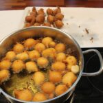 V loncu na kuhalni plošči se v vročem olju cvrejo manjši kosi testa, nekaj ocvrtih kosov je odloženih na papirnati podlagi ob loncu.